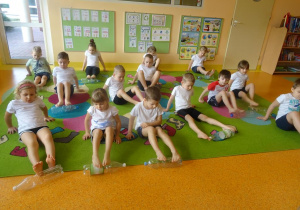 Dzieci siedzą w siadzie podpartym z ugiętymi w kolanach nogami i przesuwają stopami butelki plastikowe po dywanie.